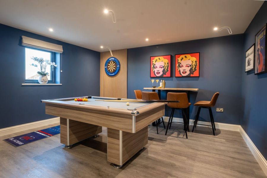 Games room at Wheatsheaf Barn, a self-catering holiday cottage, with a pool table, dartboard, bar table with stools, and framed pop-art prints on dark blue walls.