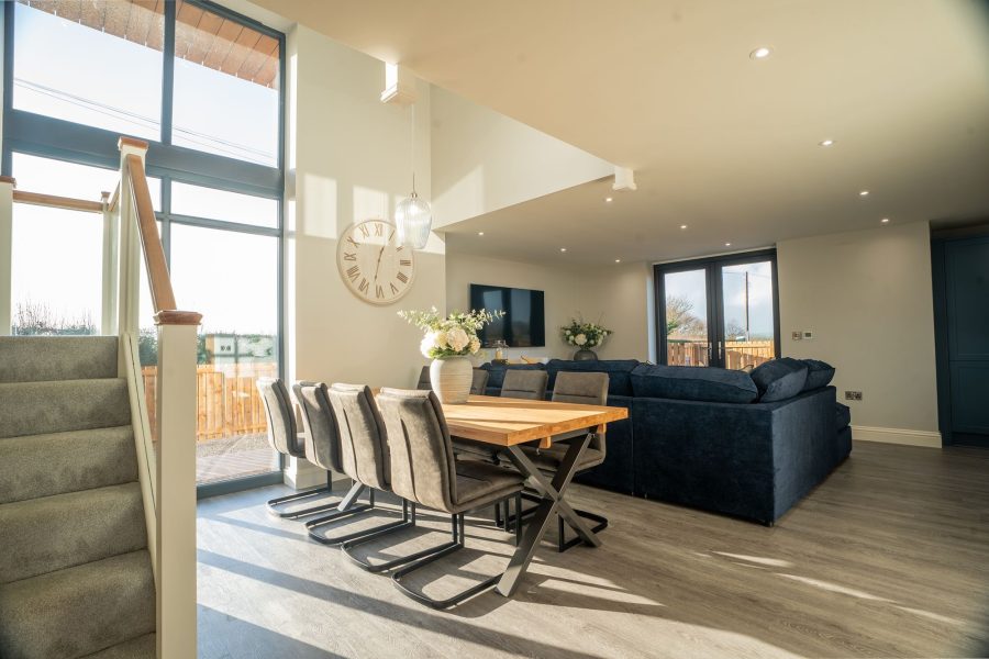 Open-plan dining and living area at Wheatsheaf Barn with dining table and chairs, blue sofa, staircase, and large windows.
