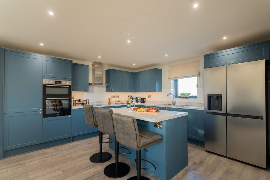 Modern self-catering kitchen at Wheatsheaf Barn with blue cabinets, central island and stools, built-in oven, and stainless steel fridge-freezer.