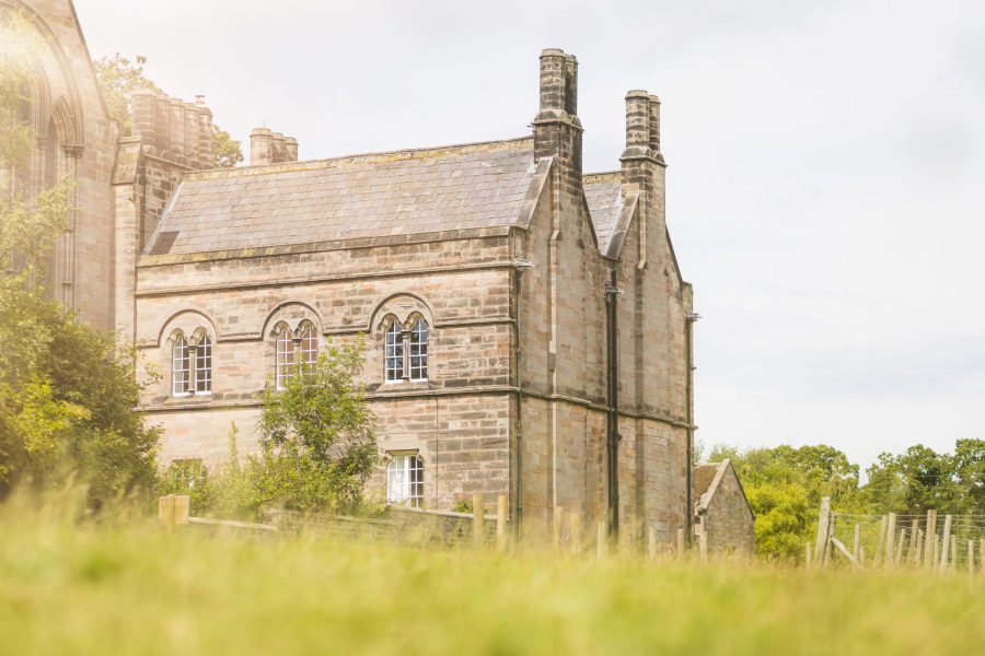 Exterior of Aquarius Rising, a historic stone building with arched windows and tall chimneys beside open green fields.
