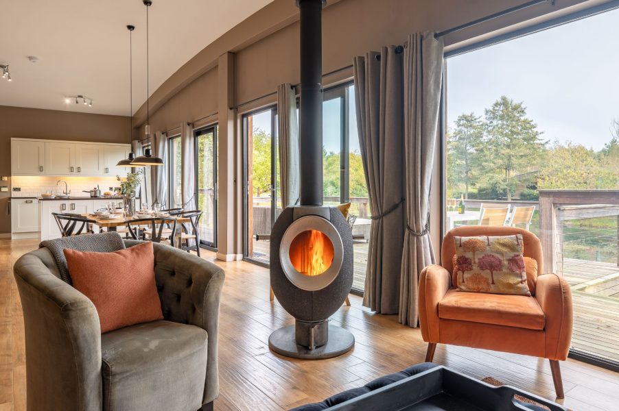 Open-plan living and dining area at Willow, featuring a central wood-burning stove and large windows opening onto the lakeside decking.