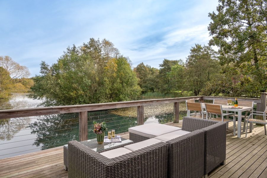 Private lakeside decking at Willow Lodge, with outdoor sofa seating, a dining table, and views across the water and surrounding trees.