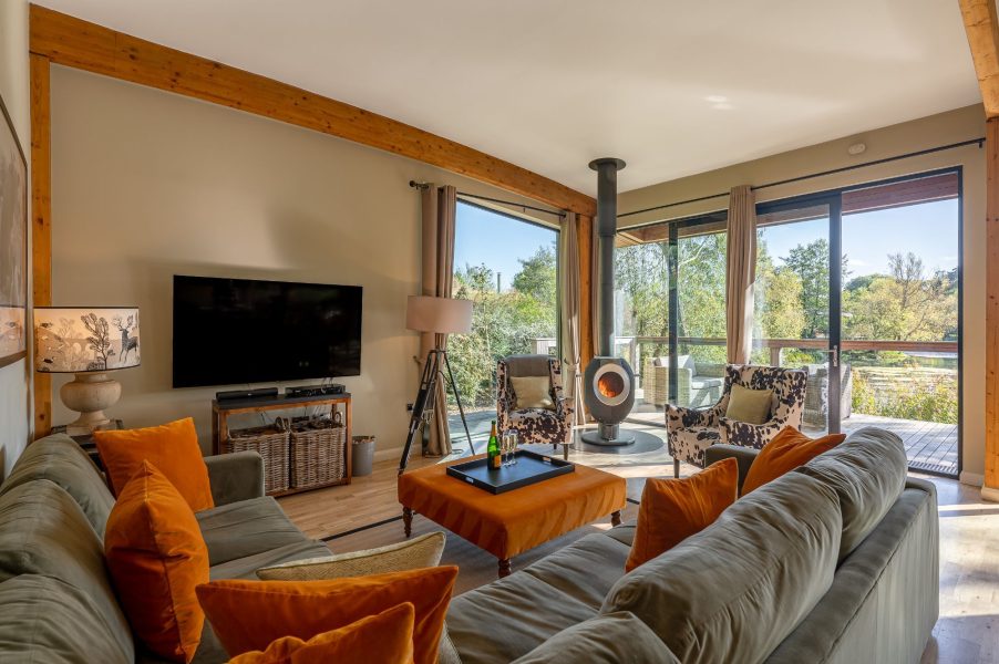 Bright, modern living room at Imogen lodge, Brompton Lakes with sofas, a wall-mounted TV, and large windows opening to a scenic outdoor view.