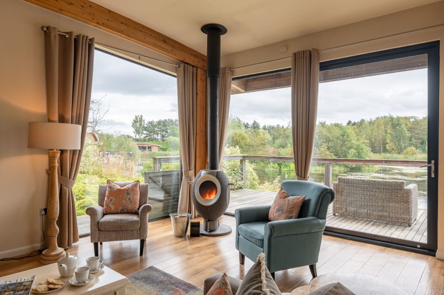 Living area in William lodge at Brompton Lakes with sofa, log burner and panoramic views of lake and surrounding nature.