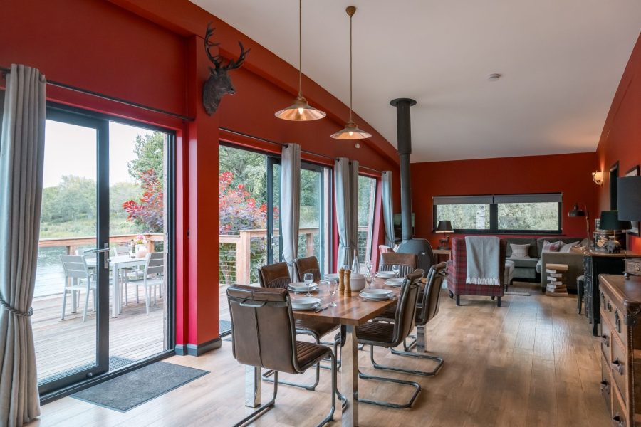 Open-plan dining and living area at Isabella with a dining table, wood-burning stove, seating area, and large windows looking out to the trees.