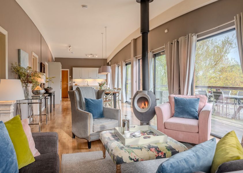 Cosy open-plan living area in Flora lodge at Brompton Lakes with log burner, armchairs and views over lakeside decking.