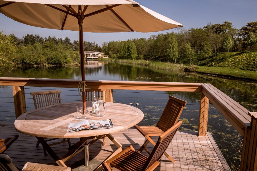 Private lakeside decking at Flora lodge at Brompton Lakes with outdoor dining table, parasol and tranquil water views.