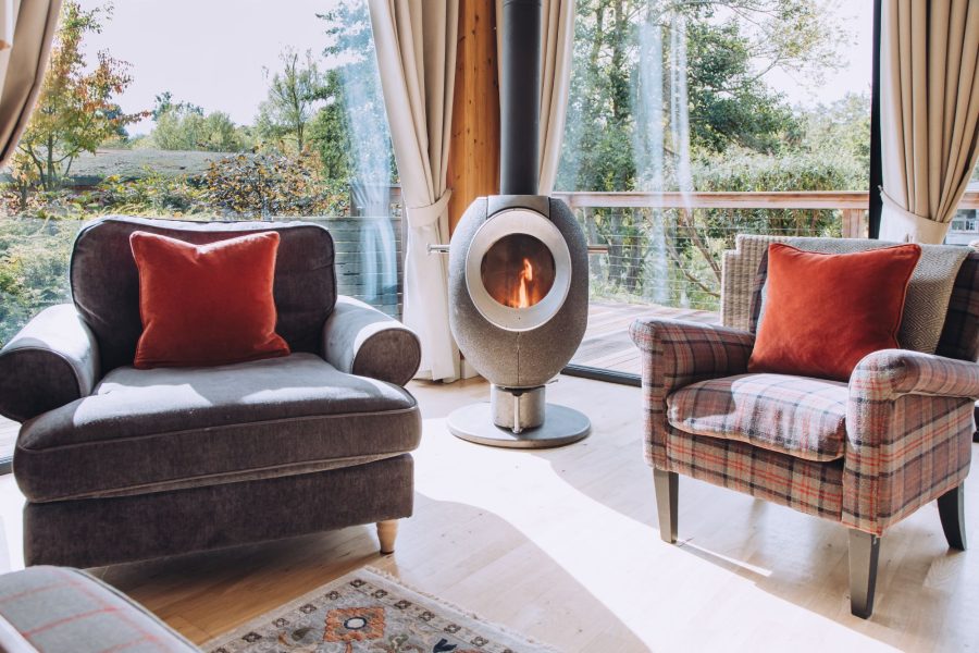 Open-plan living space at Cara lodge with wood burner, armchairs, large windows and scenic nature views at Brompton Lakes, Yorkshire.