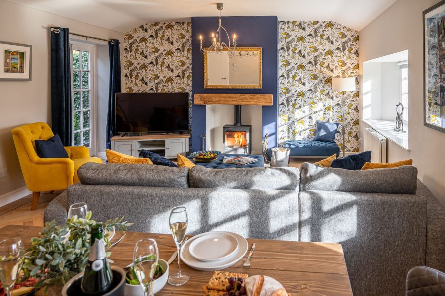 Open-plan living and dining space in The Old Potting Shed featuring a cosy sofa, wood burning stove, and vibrant patterned feature wall.