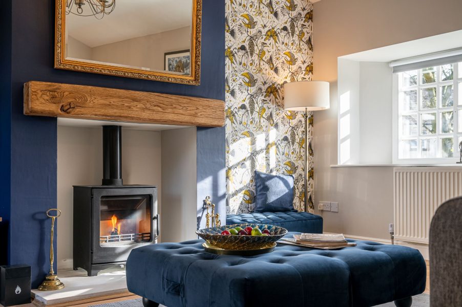 Cosy living area in The Old Potting Shed with a lit wood burning stove, plush seating, and a comfortable reading nook by the window.