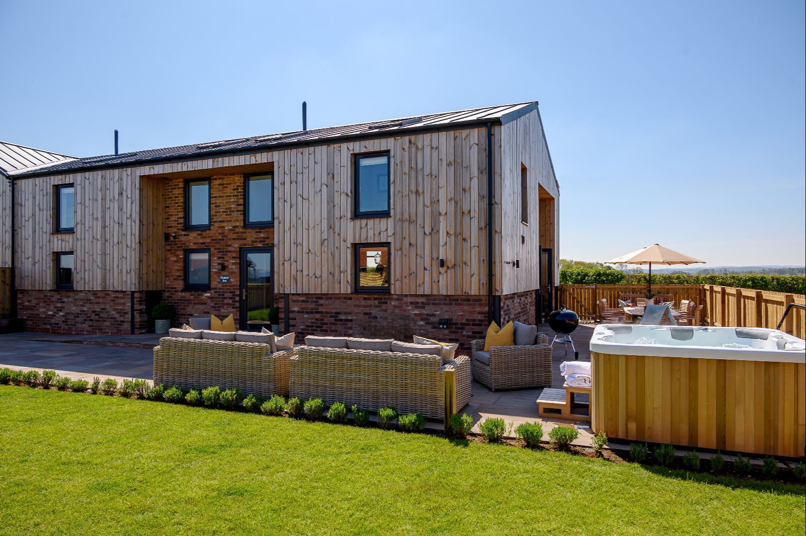 Exterior of Wheatsheaf Barn, a modern countryside holiday home with wooden cladding, patio seating, and private hot tub.