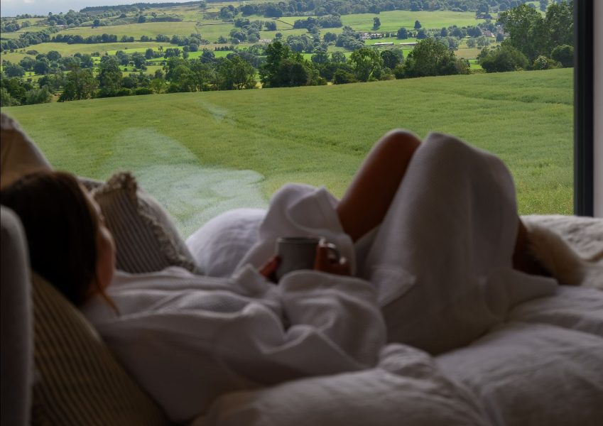 Guest relaxing in bed with a morning coffee while enjoying panoramic countryside views from a luxury lodge.