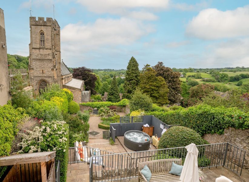 Garden and hot tub terrace at 30 Frenchgate with stone steps, planted borders, and a view towards a church tower and rolling countryside.