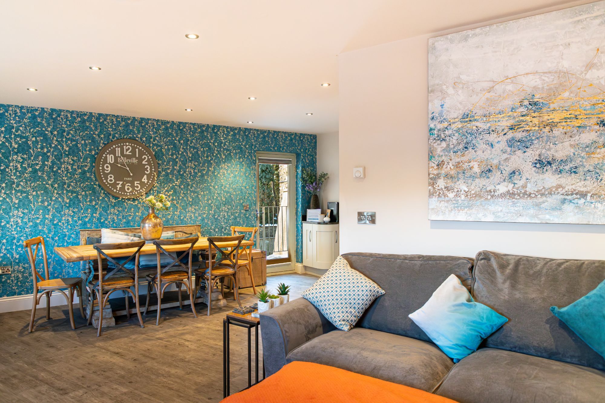 Open-plan living and dining space with wooden dining table, teal feature wall, large wall clock and comfortable sofa.