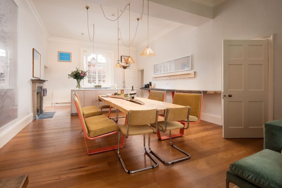 Dining area at Aquarius Rising with a wooden dining table, modern chairs, pendant lights, and a kitchen area beyond.