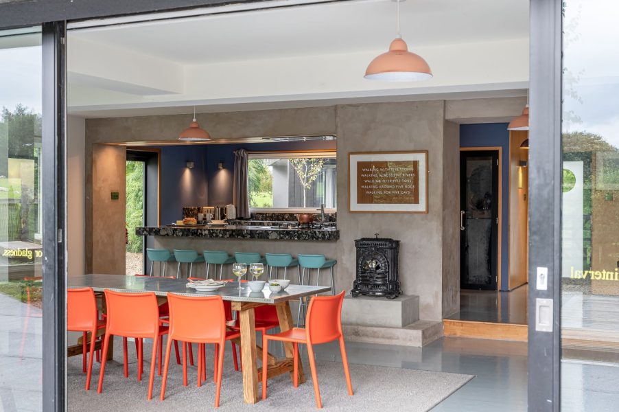 Open-plan kitchen and dining area with bright orange chairs, breakfast bar seating and large sliding glass doors to the terrace.