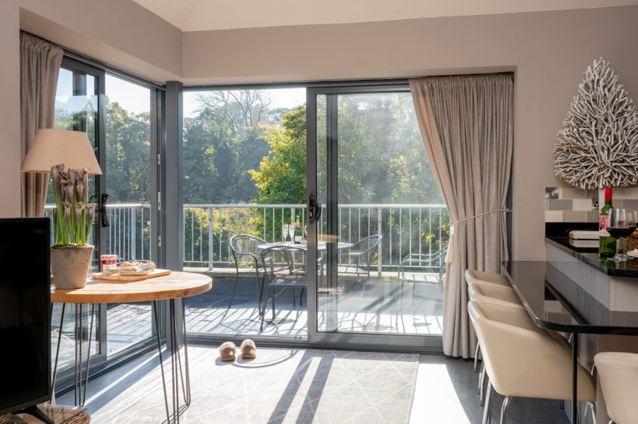 Sunlit penthouse living area with floor-to-ceiling sliding glass doors opening onto a spacious balcony, featuring outdoor seating and leafy views, alongside a stylish breakfast bar and soft neutral dÃ©cor.