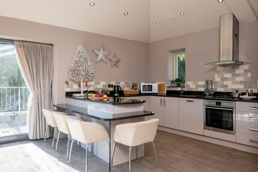 Bright, modern penthouse kitchen with a central island and breakfast bar seating, sleek cabinetry, built-in oven and extractor, and large sliding doors opening onto a balcony.