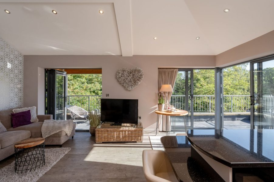Spacious open-plan penthouse living area featuring a comfortable lounge, sleek breakfast bar, and large sliding glass doors that open onto a wraparound balcony with tranquil leafy views.