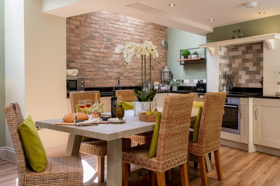 Darcy Mews dining area with a wooden table set for drinks and snacks, surrounded by wicker chairs, alongside a modern kitchen with tiled splashback and exposed brick feature wall.