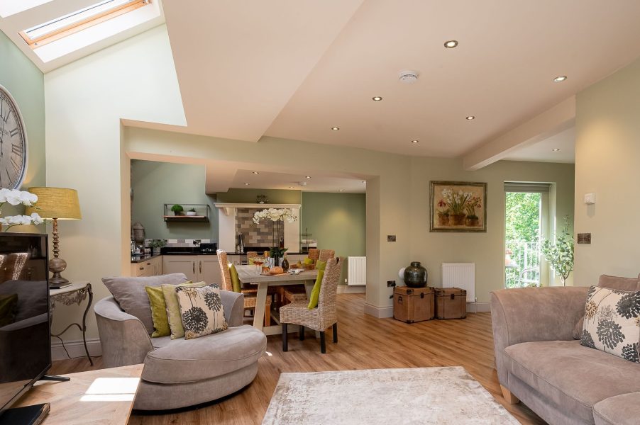 Darcy Mews open-plan living space with a sofa and armchair, neutral decor and skylight, leading through to a dining table with wicker chairs and a modern kitchen beyond.