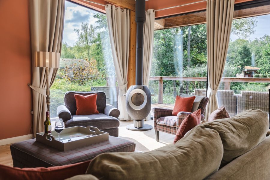 Open-plan living space at Cara lodge with wood burner, armchairs, large windows and scenic nature views at Brompton Lakes, Yorkshire.