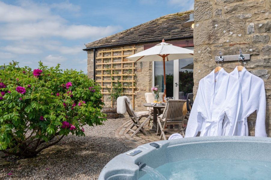 Private courtyard at Wayside Cottage featuring outdoor dining area, flowering shrubs, and a hot tub with robes ready for use.