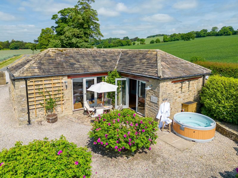 Exterior of Wayside Cottage showing a charming stone building with private patio, outdoor seating, and a hot tub set among countryside views.