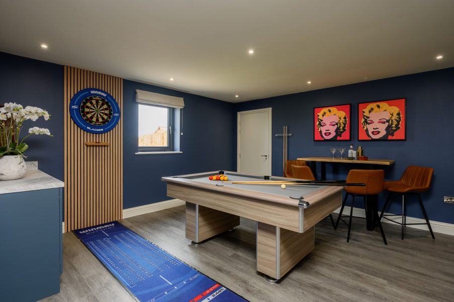 Games room at Wheatsheaf Barn, a self-catering holiday cottage, with a pool table, dartboard, bar table with stools, and framed pop-art prints on dark blue walls.